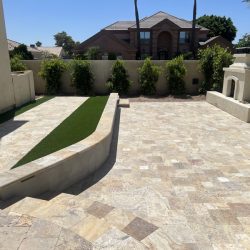 Stone-tiled backyard patio with raised planter, artificial grass strip, and outdoor fireplace against a privacy wall.