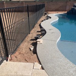 A partially installed black metal pool fence runs along the curved edge of a backyard pool on a dirt surface.