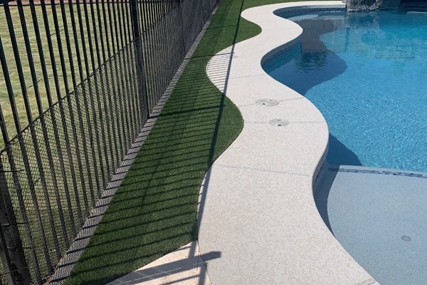 A black fence separates a grassy area from a curved pool deck beside a swimming pool in a backyard.