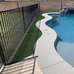 A metal fence separates a grassy area from a pool with a curved concrete border and artificial turf alongside.