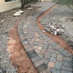 A curved stone paver walkway runs through a yard with gravel, soil, and a small pile of unused pavers on the side.