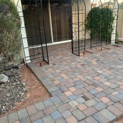A brick patio with a metal garden archway in front of a sliding glass door, bordered by gravel and plants.