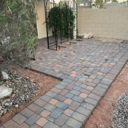 A paved patio with multicolored bricks, a metal trellis, and a small garden area beside a beige brick wall.