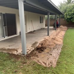 Concrete patio with soil and mud dug up along the edge; two people working near the far end under a covered area.