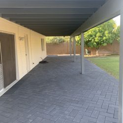Covered patio with gray herringbone pavers, adjacent to a house, facing a fenced yard with trees in the background.