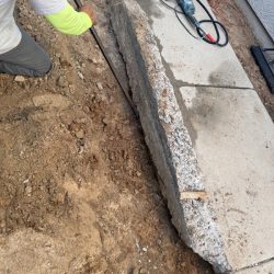 Person kneeling on dirt next to a broken concrete slab and construction tools near a building foundation.
