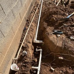 PVC pipes are installed in a trench next to a brick wall, with tools and loose wires scattered on the ground.