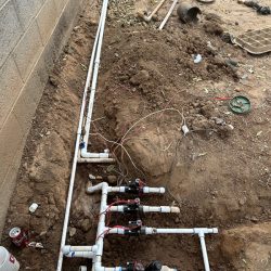 Irrigation system with PVC pipes, valves, and wiring installed in a dug trench next to a concrete wall.