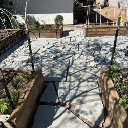 Stone pathway with irregular slabs leads through a garden with raised wooden beds and metal trellises under sunlight.