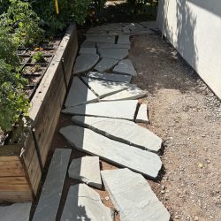 A stone pathway with irregular slabs leads beside a raised garden bed and along the side of a building.