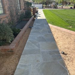 A stone walkway runs alongside a brick house, bordered by grass and a dirt area under clear daylight.