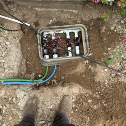Person standing near an open irrigation valve box with exposed pipes, wires, and soil in a garden area.