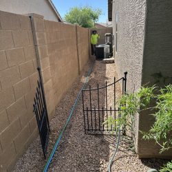 A person stands near an air conditioning unit in a narrow, rock-covered side yard with a garden hose on the ground.
