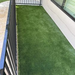 A small outdoor patio area with artificial grass flooring, bordered by a black metal railing and a beige wall with a window.