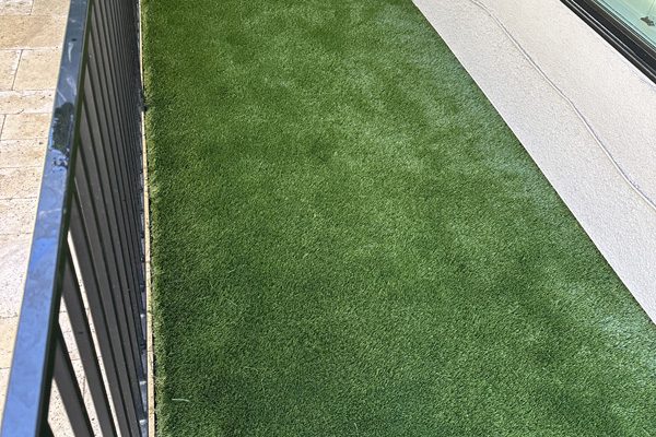 A small fenced patio area with artificial grass next to a building and large window.