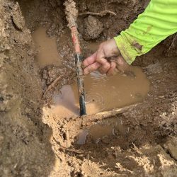 A person's hand repairs a leaking water pipe in a muddy hole filled with water.
