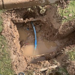 Exposed blue pipe in a muddy, water-filled hole in the ground next to a patch of grass and concrete curb.