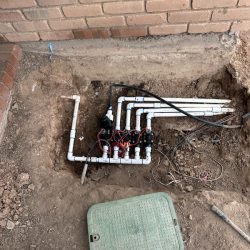 Exposed irrigation valve system with PVC pipes and wiring installed in a ground pit near a brick wall.