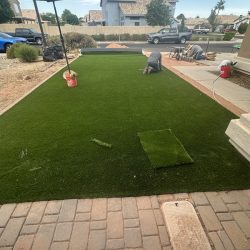 Two workers install artificial grass in a front yard, with tools and materials visible on the lawn and patio area.