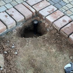A sprinkler head exposed in a hole surrounded by bricks and soil during maintenance work.