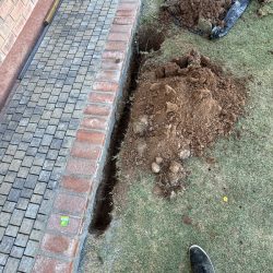 A trench and a hole dug in soil next to a brick patio, with soil piles and tools visible on the grass.