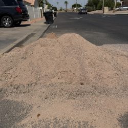 A pile of sand or gravel on a residential street blocking part of the road, with cars and houses in the background.