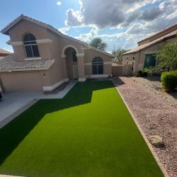 A suburban house with artificial green lawn, rock landscaping, and partly cloudy sky with sunlight overhead.