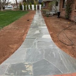 A stone walkway leads from a brick-edged entrance through a yard toward a house with trees and grass on either side.