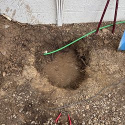 A muddy hole in the ground near a white wall with exposed pipes, a green hose, and red-handled pliers nearby.